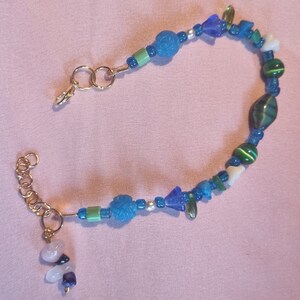 May include: A gold chain bracelet with blue, green, and white beads. The bracelet has a clasp and a charm with a white stone.