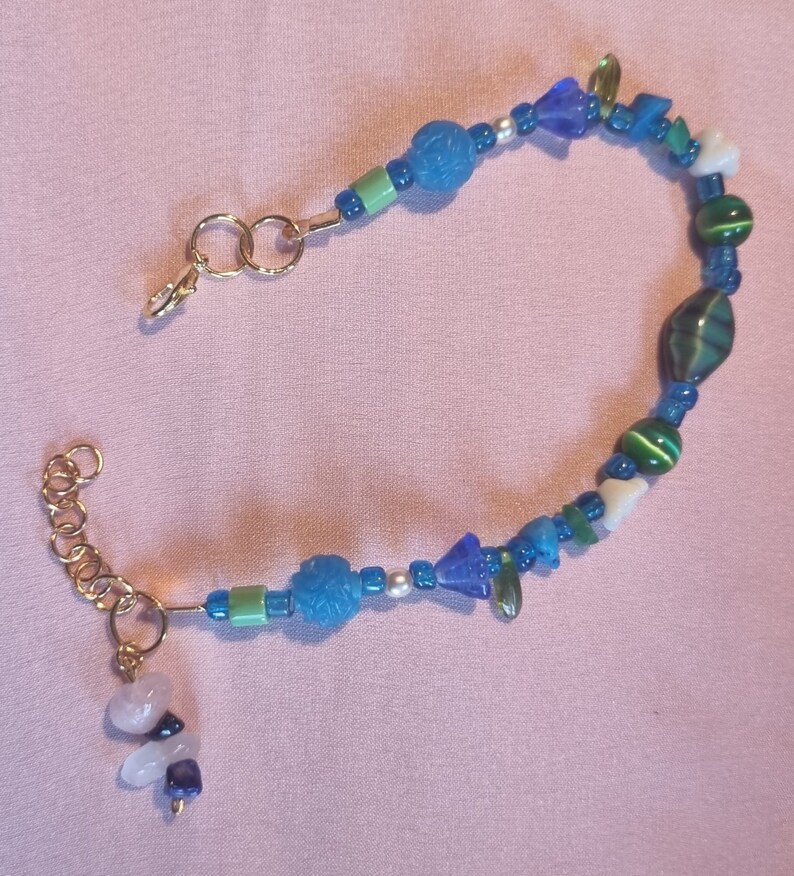 May include: A gold chain bracelet with blue, green, and white beads. The bracelet has a clasp and a charm with a white stone.