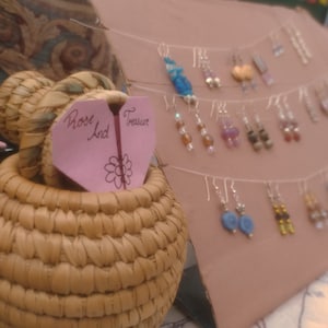 May include: A display of handmade earrings with various colours and styles. The earrings are hanging from a string and are displayed on a brown cardboard background. A pink heart-shaped sign with the text "Rose And Treasure" is in the foreground.
