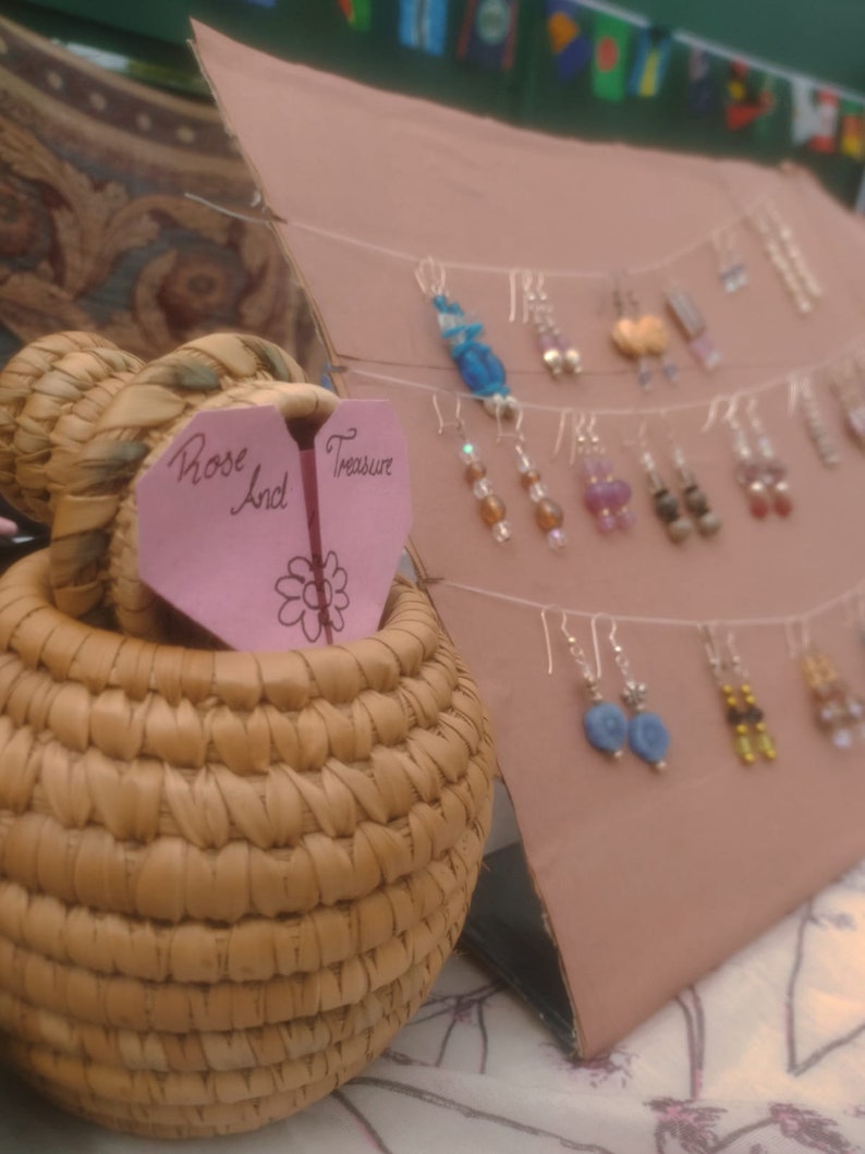 May include: A display of handmade earrings with various colours and styles. The earrings are hanging from a string and are displayed on a brown cardboard background. A pink heart-shaped sign with the text "Rose And Treasure" is in the foreground.