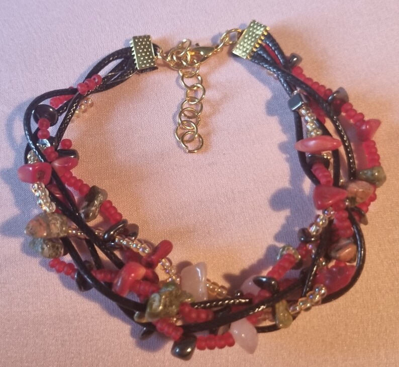 May include: Multi-strand bracelet with red, pink, green, and black beads and stones. The bracelet has a gold clasp and is made of black cord.