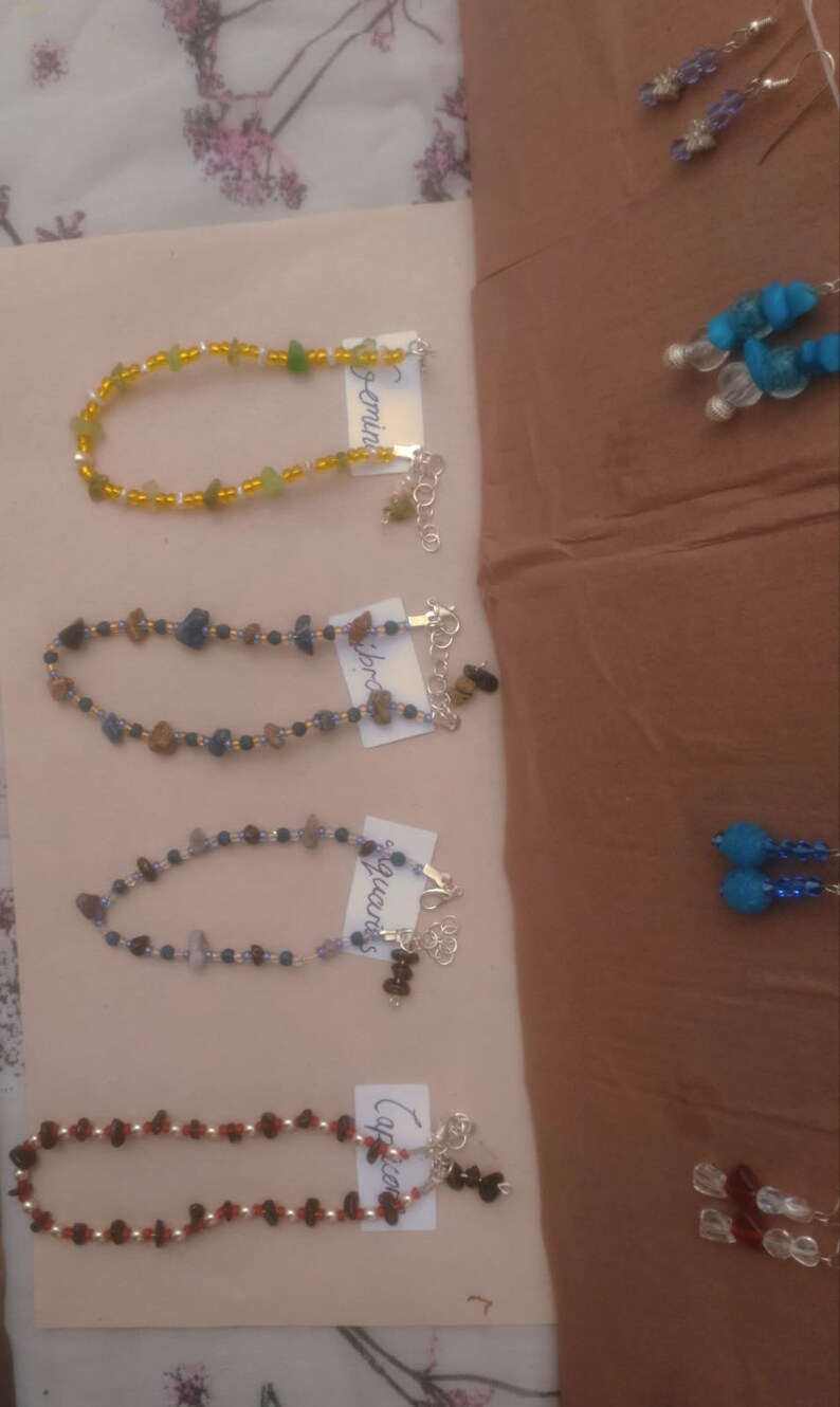May include: Four bracelets made with different colored beads and stones, each with a tag labeled with a zodiac sign: Gemini, Libra, Aquarius, and Capricorn.
