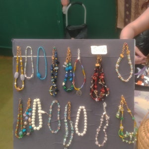 May include: A display of 12 colorful beaded bracelets with various styles and colors, including blue, green, red, white, and black. The bracelets are arranged on a gray board with the word "Sold!" written on a white tag.