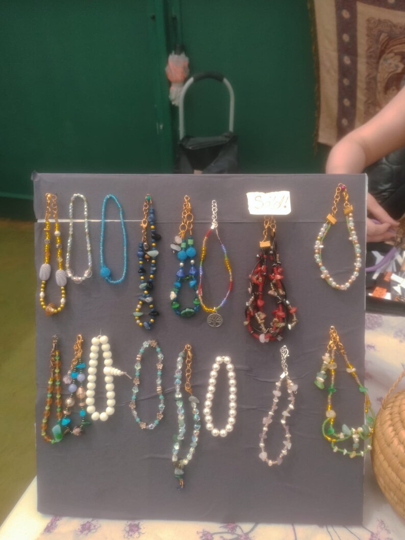 May include: A display of 12 colorful beaded bracelets with various styles and colors, including blue, green, red, white, and black. The bracelets are arranged on a gray board with the word "Sold!" written on a white tag.