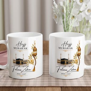 Peut inclure: Deux mugs en céramique blanche avec l'inscription "Hajj Mubarak" et "Fatima Zahra". Les mugs présentent une illustration dorée d'une mosquée, d'une lanterne et d'éléments floraux. La date 2026-1447 est également imprimée.