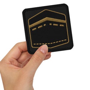 May include: Black square patch with a gold embroidered design of a building. The patch is held in a hand against a white background. The building design has a box shape with a dotted line at the bottom.
