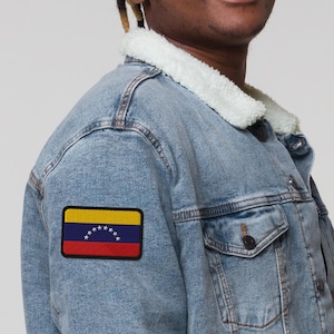 May include: Light blue denim jacket with a white faux shearling collar. The jacket features a patch of the Venezuelan flag on the left arm. The flag has yellow, blue, and red stripes with white stars.