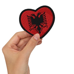 May include: A heart-shaped embroidered patch featuring a red background with a black double-headed eagle emblem in the center. The patch has a thick black stitched border around the entire edge, creating a distinct outline for the heart shape.