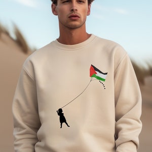 Może przedstawiać: Beżowa bluza z sylwetką dziecka latającego na latawcu z nadrukowaną flagą palestyńską. Osoba ma na sobie beżowe spodnie i stoi na piaszczystej plaży.