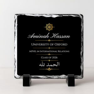 May include: A black square plaque with white and gold text, including the name Aminah Hassan, University of Oxford, and Class of 2026. The plaque has a decorative gold emblem and is supported by a black stand.
