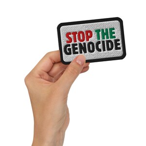 Op de afbeelding: Geborduurde patch met de tekst "STOP THE GENOCIDE". De patch heeft een rechthoekige vorm met een zwarte rand. De tekst is in rood, groen en zwart. De patch wordt door een hand vastgehouden.
