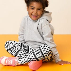 May include: A young person wearing a gray hoodie and patterned black and white leggings. Pink and white sneakers and a pink object are on the orange surface. The background is a light beige color.