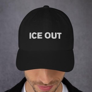 May include: Black baseball cap featuring the white text "ICE OUT". The cap has a curved peak and a structured design. The hat is worn by a person wearing a black jacket.