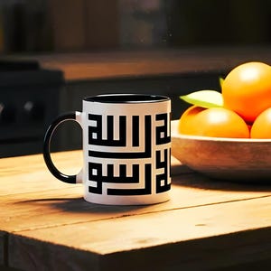 May include: A white ceramic mug with a black interior and handle, adorned with black Arabic calligraphy. The mug is placed on a wooden table, beside a wooden bowl filled with oranges. The background shows a blurred kitchen.