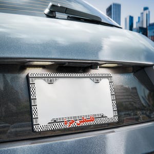 May include: A license plate frame with a black and white checkered pattern and the words "Palestine Libre" in red Arabic script. The frame is mounted on the back of a gray vehicle.