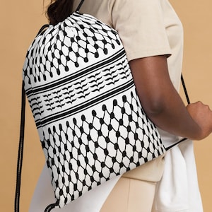 Puede incluir: Una mochila blanca con cordón y un patrón geométrico negro. La bolsa presenta un patrón repetitivo de puntos y líneas negras interconectadas, con rayas negras horizontales y un diseño decorativo. La bolsa es llevada por una persona.