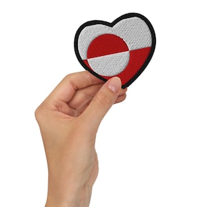 May include: Heart-shaped embroidered patch featuring the flag of Greenland. The patch has a black border, with a white and red design. The patch is held in a hand.