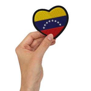 May include: Heart-shaped embroidered patch featuring the flag of Venezuela. The patch has a yellow top, a blue center with white stars, and a red bottom. The patch is held in a hand against a white background.
