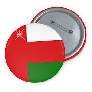 May include: Two round pinback buttons. One button displays the flag of Oman, featuring red, white, and green colors with a white emblem. The other button shows the silver back with a pin.