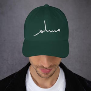 Op de afbeelding: Een donkergroene baseballcap met het wit geborduurde woord "Muslim" in Arabisch schrift. De cap heeft een gebogen klep en een klassiek zes-panelen ontwerp. Gedragen door een persoon.