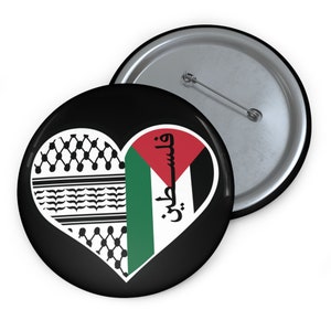 Könnte beinhalten: Ein schwarzer Button mit einem herzförmigen Design, das die palästinensische Flagge und das traditionelle Keffiyeh-Muster zeigt. Das Herz enthält die Farben Rot, Weiß, Grün und Schwarz, mit arabischem Text. Ein zweiter Button ist sichtbar.