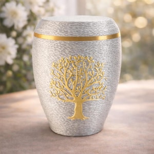May include: Silver urn with a gold band and a gold tree design. The urn has a textured surface and a rounded shape. The background is blurred with white flowers.