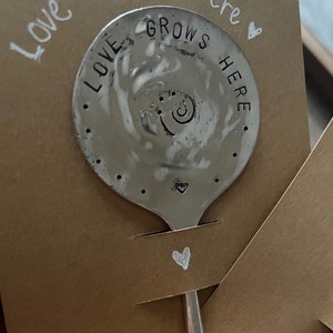May include: A silver spoon with the words "Love Grows Here" stamped on it. The spoon is on a brown card with the same words written in white marker.