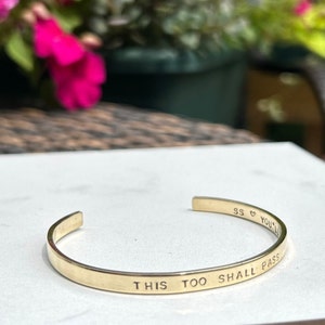 May include: A gold-colored cuff bracelet with the engraved phrase "THIS TOO SHALL PASS." The bracelet has rounded edges and a simple, elegant design. The bracelet is on a white surface.