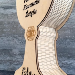Pode incluir: Recordação em madeira em forma de cálice com o texto "Pierwsza Komunia Święta" e o nome "Fabian" com a data 16.07.2026. O cálice tem um emblema IHS decorativo e um design em camadas.