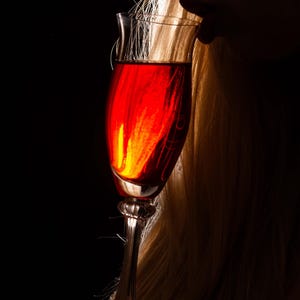 Könnte beinhalten: Ein schlankes Weinglas, gefüllt mit einer tiefroten Flüssigkeit, möglicherweise Wein. Das Glas wird in der Nähe des Gesichts einer Person gehalten, mit langem blondem Haar. Der Hintergrund ist tiefschwarz.
