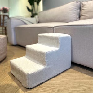 Puede incluir: Un escalón para mascotas de tres escalones en color blanco roto neutro. Los escalones están hechos de un material suave y texturizado, y están diseñados para ayudar a las mascotas a alcanzar superficies más altas. El escalón está colocado sobre un suelo de madera junto a un sofá beige.