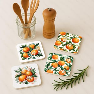 Könnte beinhalten: Vier quadratische Untersetzer mit einem Aquarelldesign aus Orangen, grünen Blättern und weißen Blüten. Holzutensilien in einem Glas, eine hölzerne Pfeffermühle und ein Rosmarinzweig sind ebenfalls vorhanden.