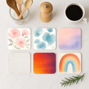 Puede incluir: Seis posavasos cuadrados con diferentes diseños de acuarela. Los diseños incluyen flores rosas, nubes azules, un degradado de rosa y morado, un degradado de naranja y rojo, puntos blancos sobre un fondo beige y un arcoíris.