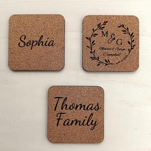 A close-up of three square cork coasters, each engraved with different personalized text (e.g., names and family monograms). The coasters are displayed on a light background, illustrating the option to customize the backside with any desired text.