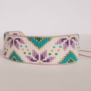 May include: A white beaded bracelet with a geometric pattern in shades of purple, teal, and gold. The bracelet has a brown leather cord.