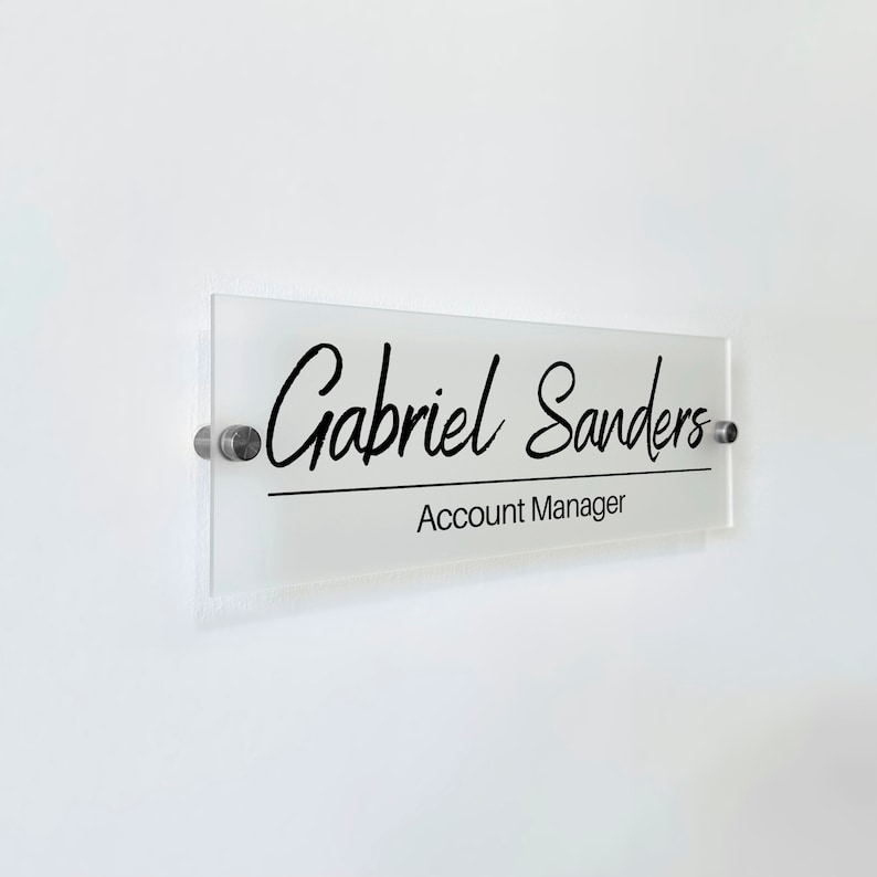 May include: A rectangular frosted acrylic office sign featuring the black printed text Gabriel Sanders in a script font and Account Manager in a sans-serif font below a horizontal line. The sign is mounted with two silver metal standoffs.