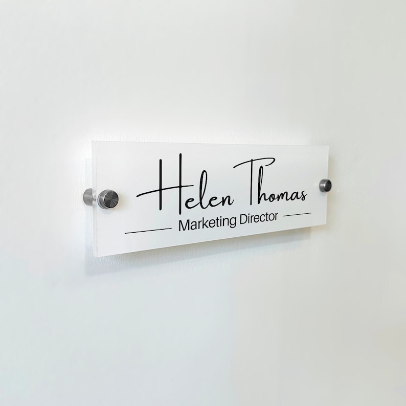 May include: A rectangular clear acrylic office sign featuring the black printed text Helen Thomas and Marketing Director. The sign is mounted to the wall with two circular silver metal standoffs on the left and right sides of the plaque.