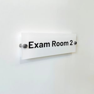 May include: A rectangular white sign with the black text Exam Room 2 printed on the front. The sign is made of a clear acrylic material and is mounted to the wall using two circular silver metal standoffs on the left and right sides.
