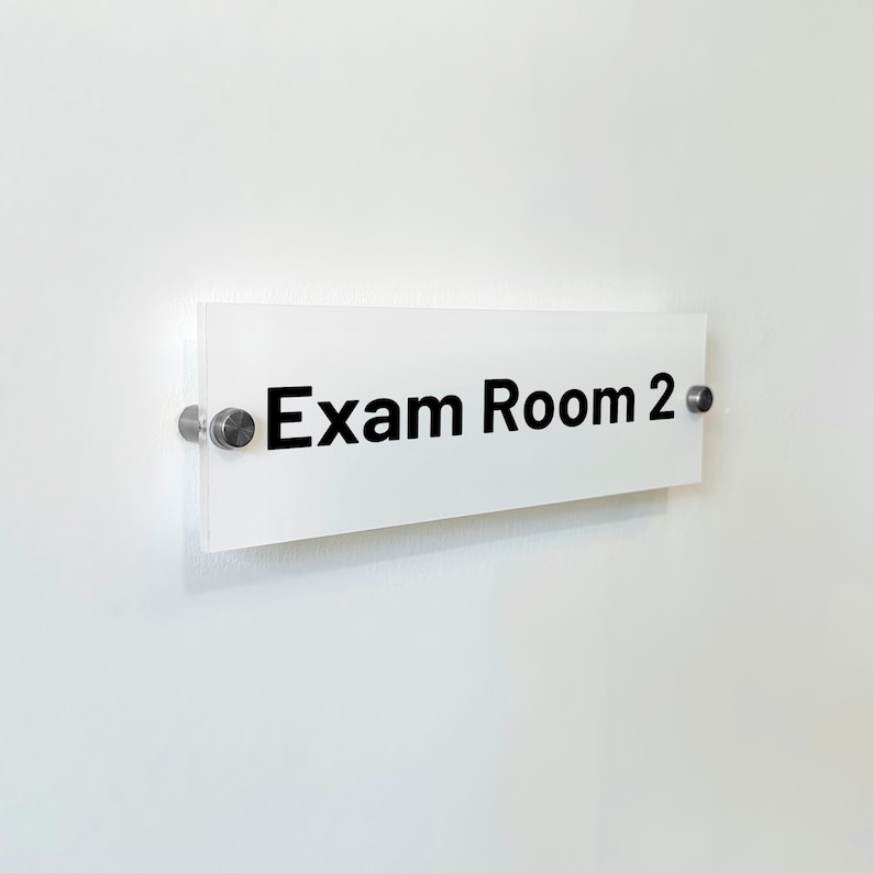 May include: A rectangular white sign with the black text Exam Room 2 printed on the front. The sign is made of a clear acrylic material and is mounted to the wall using two circular silver metal standoffs on the left and right sides.