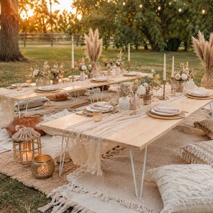 May include: Two low-profile wooden picnic tables with white hairpin legs are arranged on layered rugs and cushions. The tables are set with white plates, glassware, candles, and floral arrangements, creating a cohesive outdoor dining setup.
