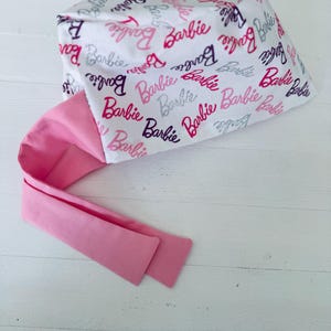 May include: A white scrub hat with a pink tie. The hat is covered in a repeating pattern of the word "Barbie" in pink, purple, and silver glitter.