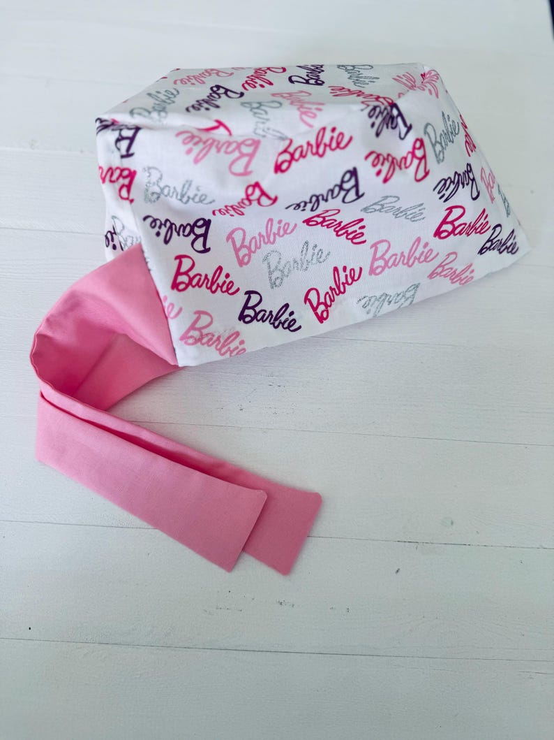 May include: A white scrub hat with a pink tie. The hat is covered in a repeating pattern of the word "Barbie" in pink, purple, and silver glitter.