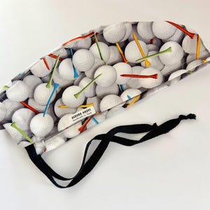 May include: A patterned surgical scrub cap featuring a golf ball design with colorful tees. The cap has a black tie closure and a tag that reads "SCRUBB AHOY!". The background is white.
