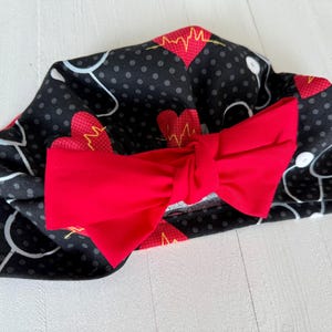 May include: A black medical scrub cap with a red bow. The cap features a pattern of red hearts, stethoscopes, and gray polka dots. The bow is a solid, vibrant red.