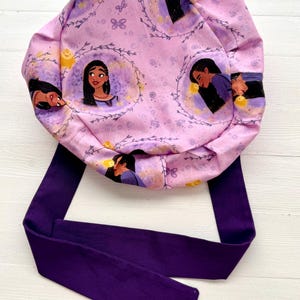 May include: A purple and pink fabric scrub hat with a tie. The hat features a repeating pattern of a cartoon woman with long dark hair and a yellow glow around her.