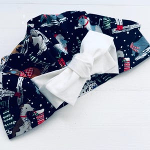 May include: A navy blue baby headband with a white bow. The headband is patterned with dogs, Christmas trees, and gifts. The bow is made of a soft, white fabric. The headband is likely for a baby or toddler.