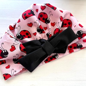 May include: A pink fabric head covering with a black bow. The fabric is patterned with red ladybugs and hearts. The ladybugs have black spots and antennae. The bow is centered on the front of the head covering.