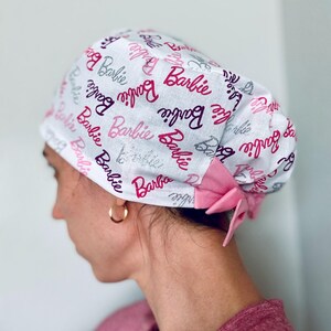 May include: A white surgical scrub hat with a pink bow and a repeating pattern of the word "Barbie" in pink, purple, and silver glitter.