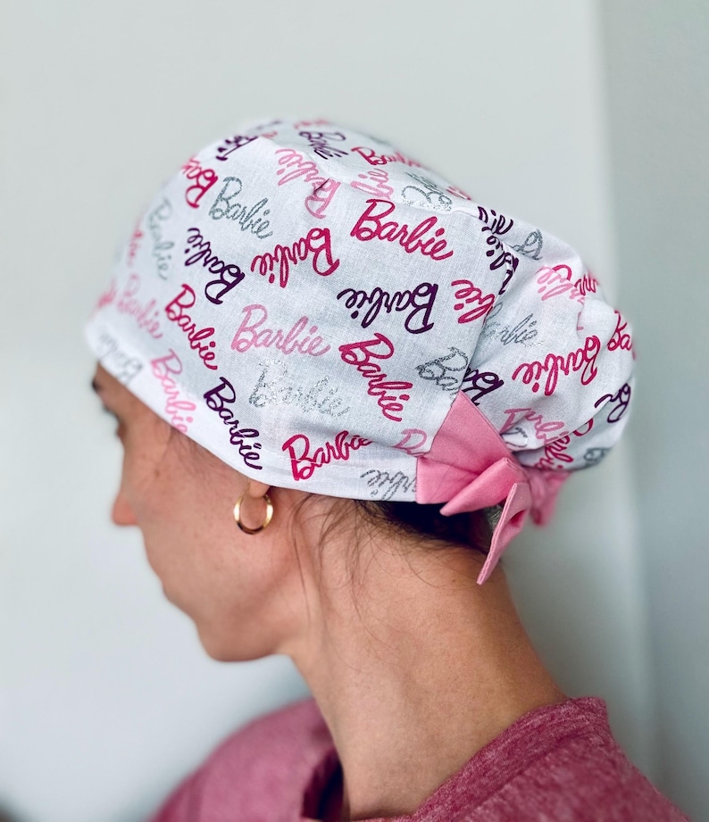 May include: A white surgical scrub hat with a pink bow and a repeating pattern of the word "Barbie" in pink, purple, and silver glitter.