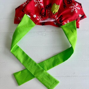 May include: A red fabric scrub cap with a green tie. The cap features white snowflakes and the text "Merry Grinchmas" with a cartoon Grinch face. The tie is a bright green color.